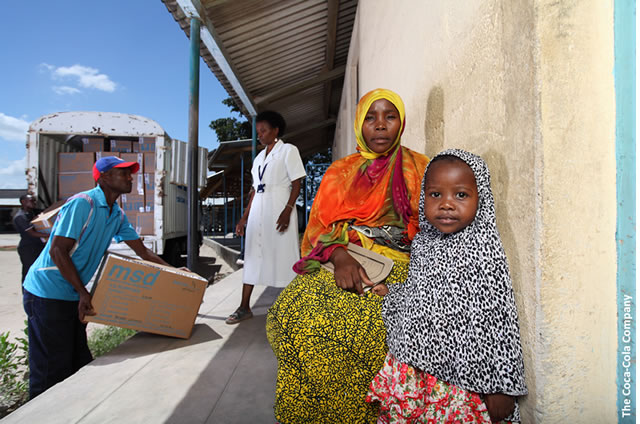A woman and child, sit on a porch where medical supplies are being delivered. Photo credit: The Coca-Cola Company