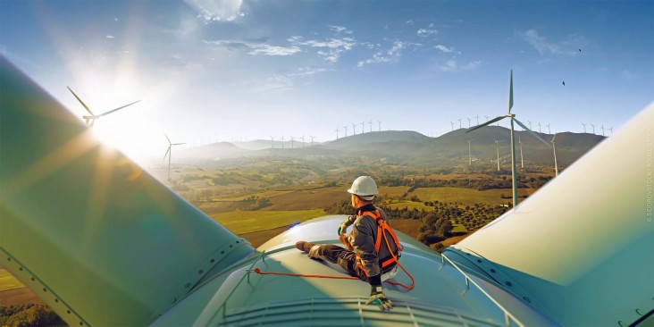 An engineer rests atop a large wind turbine admiring his work and the beautiful sunset landscape.