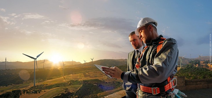 A businessman in a suit and an engineer wearing electrical and climbing safety gear look at a tablet computer while surveying a beautiful agricultural landscape in Ukraine that is dotted with wind turbines.