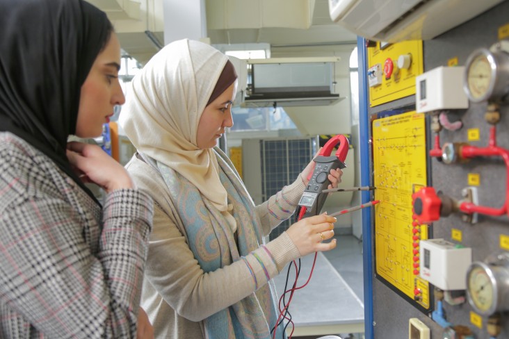 Two women stand in front a machine, looking at an electrical meter.